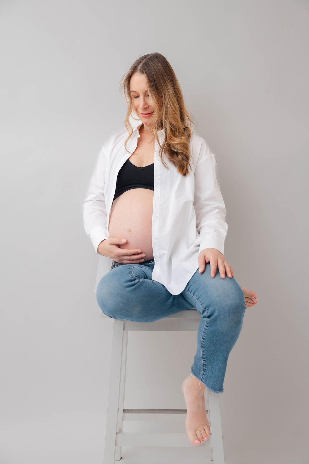 A mother to be sits on a stool in a studio in jeans and white shirt smiling down to her bump