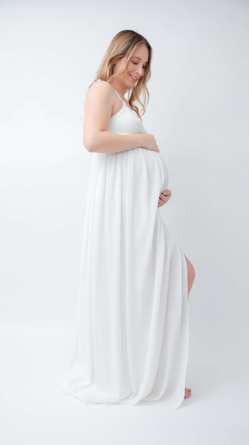 A happy pregnant woman stands in a white gown smiling down to her bump in a studio after some prenatal yoga in Torrance