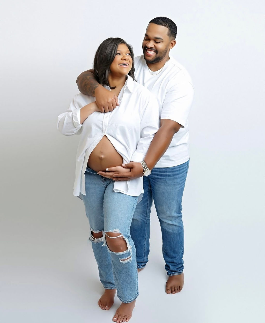A happy expecting couple stand in a studio in white shirts and jeans holding the bump after some prenatal yoga in Torrance