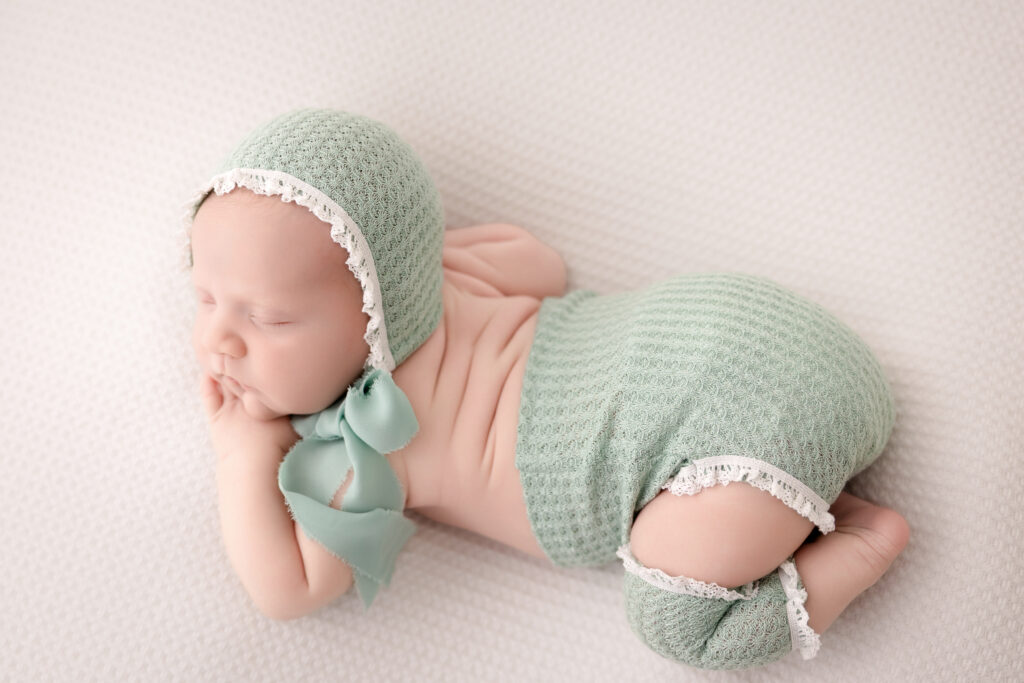 9 day old baby girl posed sleeping on belly wearing a green and lace bonnet, bottom, and leggings in studio in Redondo Beach, CA.
