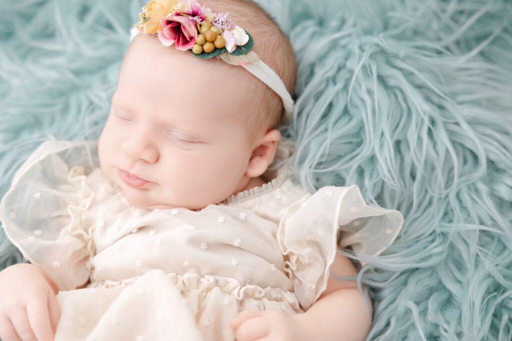 Baby girl asleep posed  against a mint green sherpa fur and wearing a cream-laced dress.