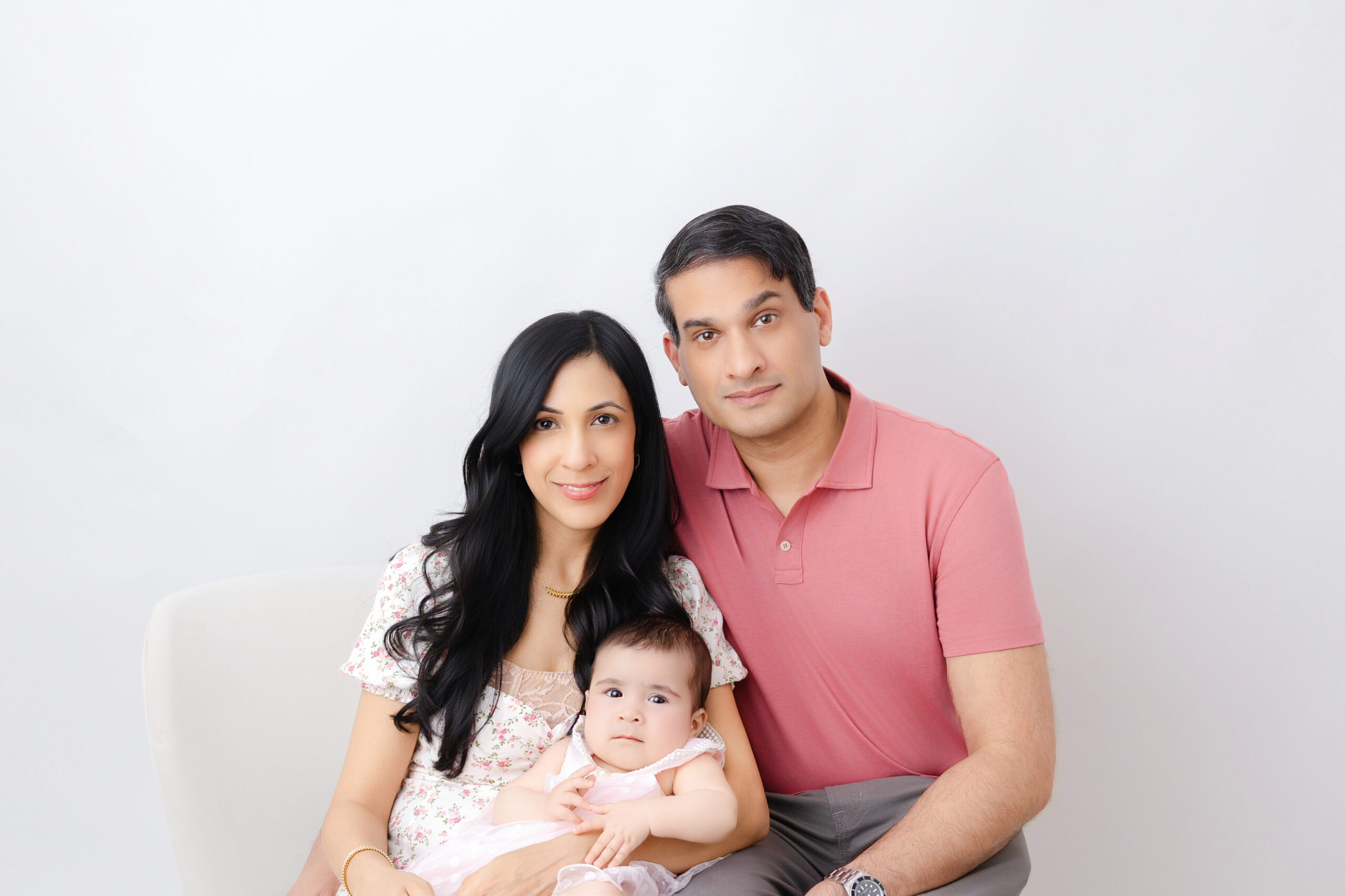 Father, mother, and 6 month old baby girl wearing different shades of pink and posed for a family portrait session at Heather Nicole Photography Studio