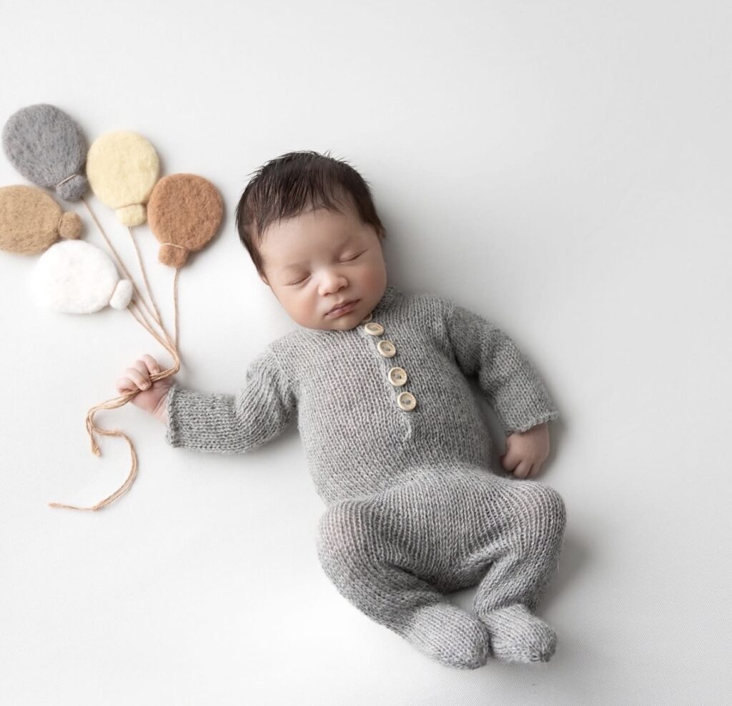 Newborn baby boy posed on his back and holding strings to 4 felt balloons.